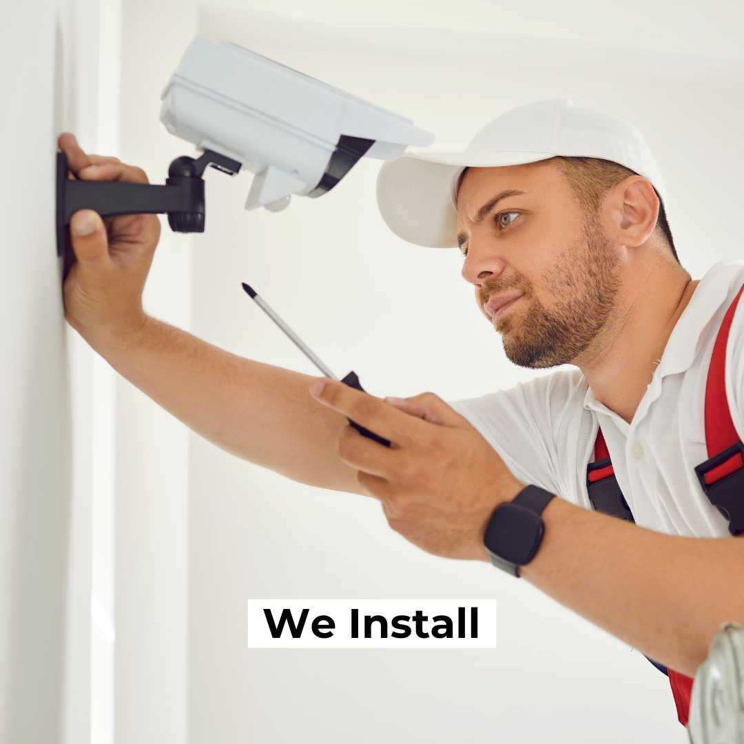 A focused technician in a white hard hat and red suspenders installs a white CCTV camera on a wall. He uses a screwdriver while attentively adjusting the camera. The bottom of the image has the text "We Install."