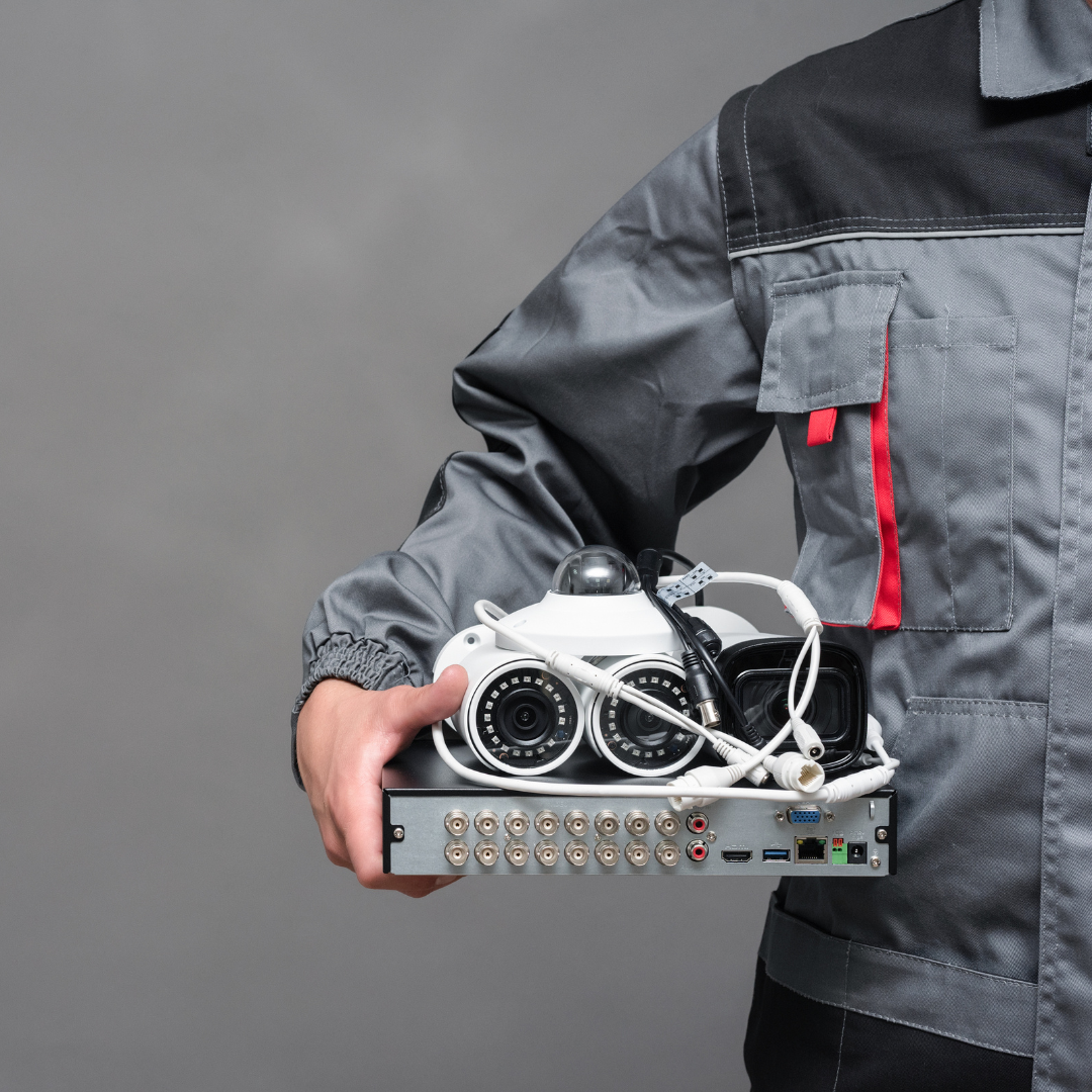 A person in a dark gray work uniform with a red accent on the pocket holds a DVR system with multiple ports, alongside two white CCTV cameras with cables. The background is a plain gray, emphasizing the equipment in the technician's hand.