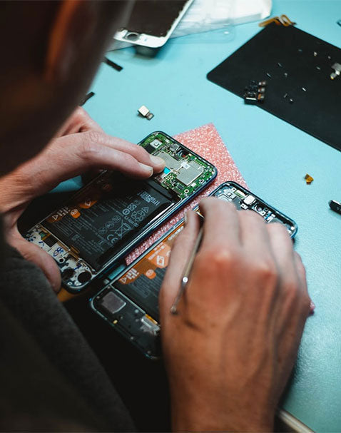 The image captures an individual working on a disassembled smartphone, possibly performing a repair or inspection. The phone's internal components, such as the battery and motherboard, are visible. The workspace has a blue surface, and there are some small parts and tools scattered around.