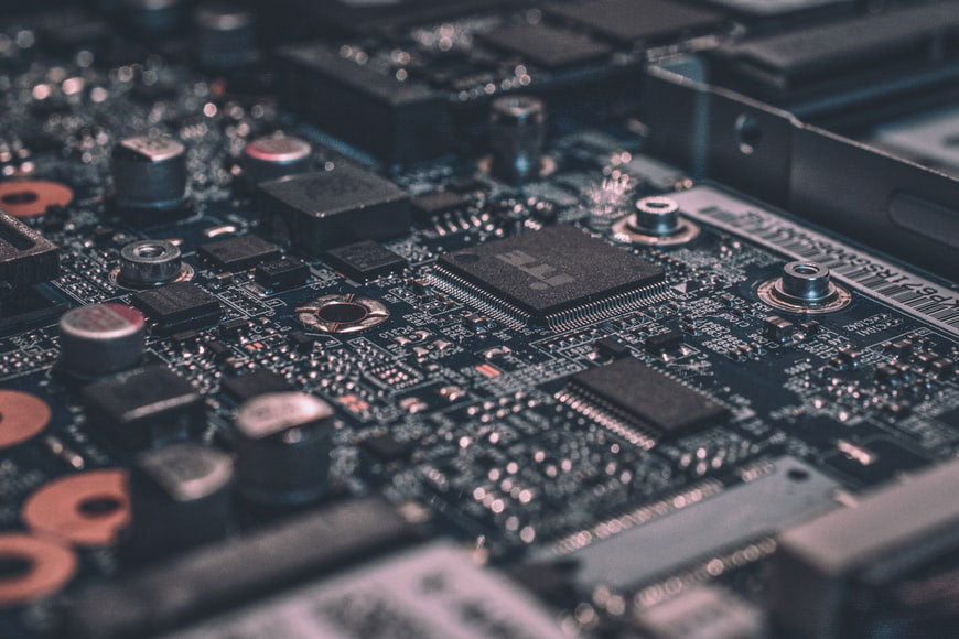 A close-up view of a complex circuit board, showcasing intricate details of electronic components, including capacitors, chips, and connectors. The board has a variety of elements in varying sizes, reflecting the intricate design and dense assembly of modern electronics. The photo captures the precise and detailed nature of technology.